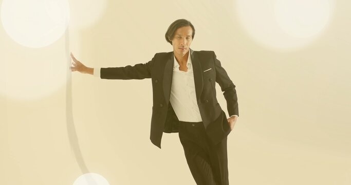 Leaning Asian model wearing black blazer and trousers extending left arm in studio, bokeh lights