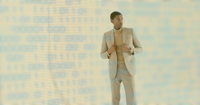 Standing man wearing tan suit and camel turtleneck, checking silver watch in studio, binary overlay