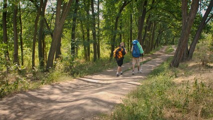 Obraz premium Two hikers with backpacks walking on a sunlit forest path surrounded by lush green trees. Vacation concept on a sunny summer day.