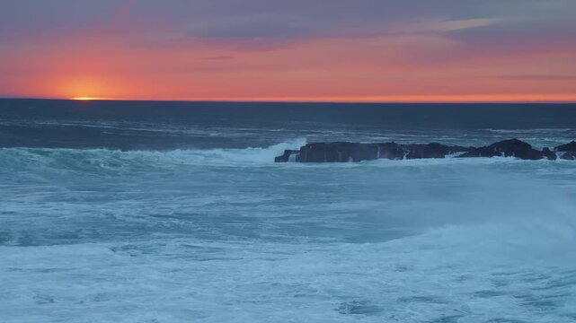 Dramatic footage of powerful ocean waves crashing against a rocky shoreline during a vibrant sunset. The sky transitions from orange to purple and blue hues, creating a serene yet powerful natural sce