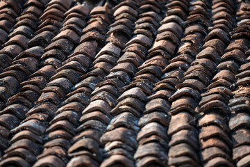 A close-up view of an old, weathered clay tile roof. The reddish-brown tiles are unevenly stacked, showing signs of age, wear, and moss or lichen growth.