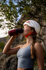 Woman in climbing gear drinking from thermal cup during mountain hike, focus on hydration and energy in outdoor sports, female alpinist resting on natural terrain