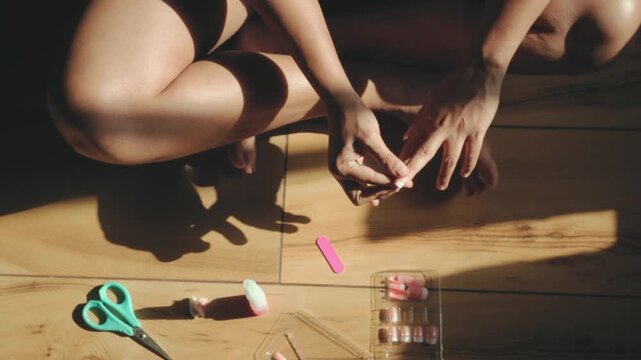Woman at home performing a self-care routine, applying false nails to her toes on a warm wooden floor, engaging in a personal moment of beauty and relaxation