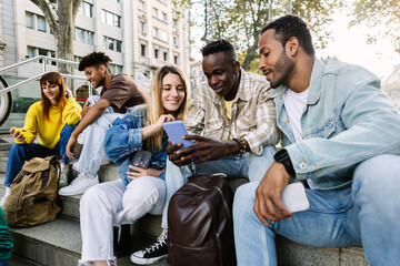 Multiracial group of millennial student friends sitting together outdoors, watching social media content on a smart phone and laughing joyfully. Youth and digital communication lifestyle concept.