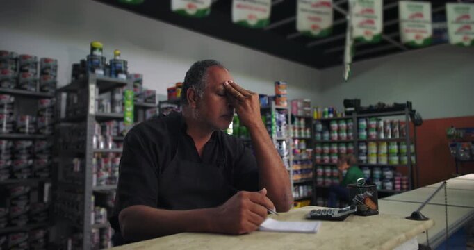 Male shop owner holding his head in frustration, overwhelmed by challenges, contemplating solutions, small business struggles in retail store environment