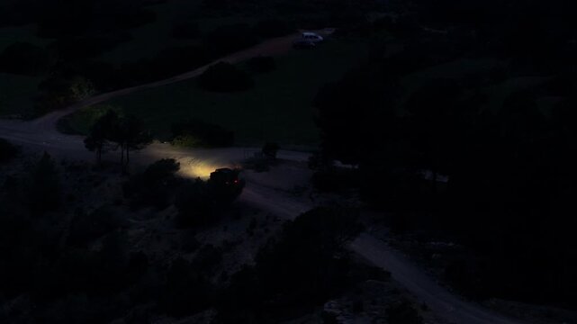 High angle tripod view of dark SUV with headlights on, driving along winding dirt road through dark rural landscape during night