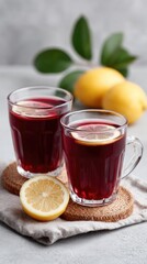 Two Glasses Of Red Fruit Drink With Lemon Slices And Whole Lemons On A Grey Textured Background With Green Leaves Studio Lighting