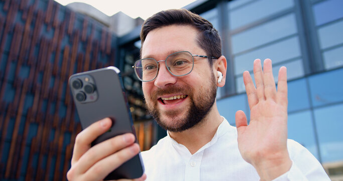 Close up of smiling Caucasian businessman in white shirt talking on video call using phone standing on street near an office building. Entrepreneur communicates remotely looking at camera. Online chat