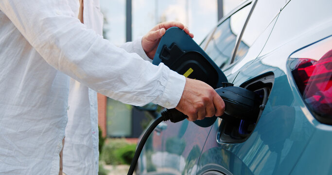 Close-up of male hands connecting an electric car charger to a discharged vehicle, starting the charging process outdoors.
