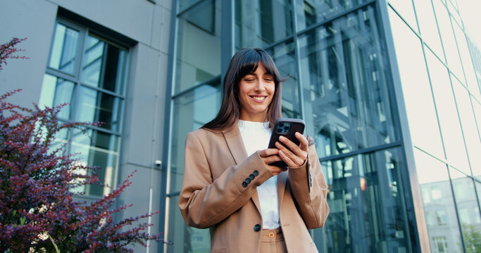 Young Caucasian businesswoman standing outside and smiling while using smartphone, with modern glass office building in the background