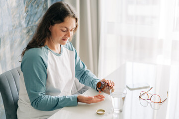 A brunette at a bright home table pours pills into her hand, and a glass of water stands next to...