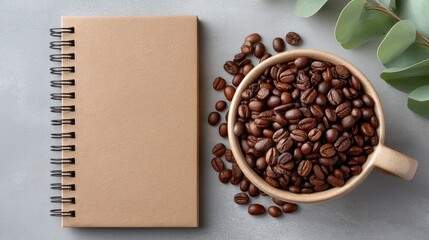 Overhead view of roasted coffee beans spilling from a light brown mug next to a closed spiral bound notebook on a textured gray surface with eucalyptus leaves on the side
