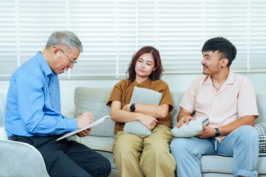 Asian couple sitting close during counseling session with male psychiatrist taking notes, woman holding cushion looking emotional while man listening beside, concept relationship therapy, empathy