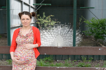 A pregnant girl in a dress and a red blouse stands near a cage with a peacock