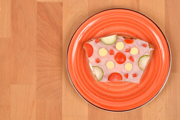 An overhead shot shows a slice of bread topped with ham, cheese, cucumber, and tomato, served on a textured orange plate centered on a wooden table