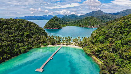 Explore the tranquil waters of Ko Ngam Beach, surrounded by lush greenery and distant mountains. Relax on the sandy shore as boats drift lazily in the turquoise sea under a bright blue sky.