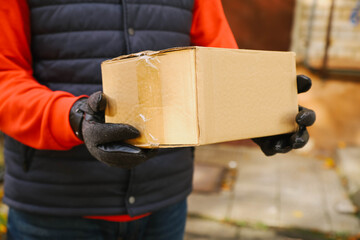 A person wearing a black vest and orange sweater holds a cardboard box with both hands.