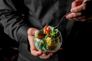 A chef's hands hold a glass bowl of fresh green salad with croutons and tomatoes, preparing to serve it with a fork. Close-up, dark background
