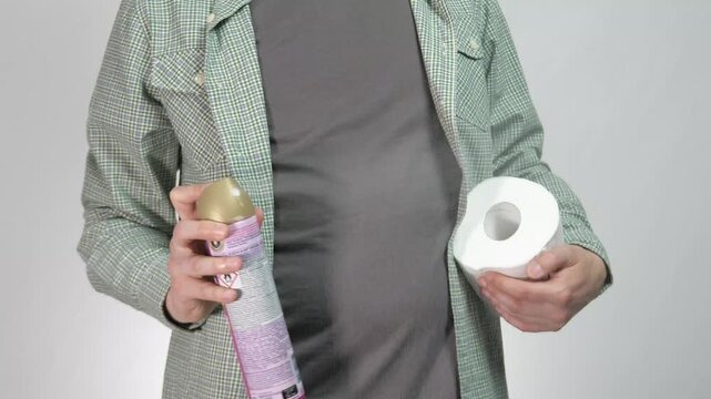 A close up shot of a persons hands performing a routine cleaning task. One hand holds a fresh roll of toilet paper while the other sprays air freshener, representing bathroom maintenance and creating