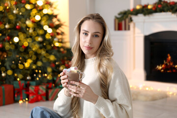 Beautiful woman with tasty hot cocoa by fireplace in room decorated for Christmas