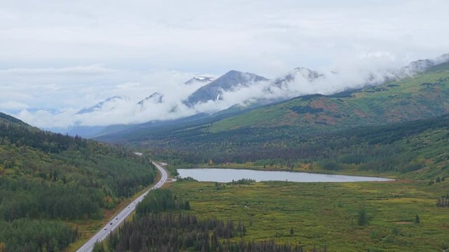Aerial view of a winding road cutting through a lush green valley with a lake and mountains shrouded in mist, Seward, Alaska, United States.