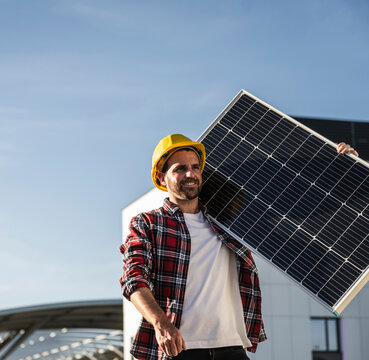 Smiling engineer wearing hardhat carrying solar panel on shoulder at terrace