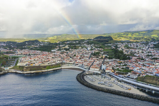 Aerial view of a vibrant town nestled by the sea, a rainbow arches above the landscape, painting a breathtaking panorama, Angra do Hero&Atilde;&shy;smo, Azores, Portugal.