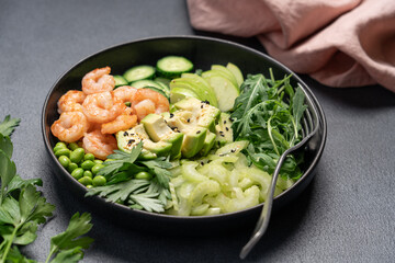 Healthy green shrimp salad with avocado, celery, apple, arugula, edamame beans, cucumber, and parsley in black bowl on dark background, top view