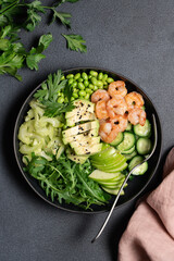 Healthy green shrimp salad with avocado, celery, apple, arugula, edamame beans, cucumber, and parsley in black bowl on dark background, top view