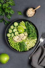 Healthy green salad with avocado, celery, apple, arugula, edamame beans, cucumber, and parsley in black bowl on dark background, top view