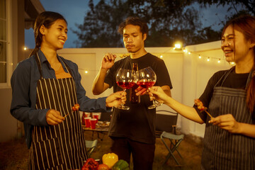 An Asian woman and friends toast with wine during a cozy backyard BBQ. Grilling skewers over charcoal under string lights, they celebrate friendship, joy,  togetherness in a festive outdoor evening