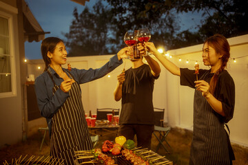 An Asian woman and friends toast with wine during a cozy backyard BBQ. Grilling skewers over charcoal under string lights, they celebrate friendship, joy,  togetherness in a festive outdoor evening