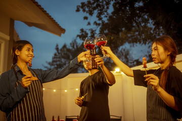 An Asian woman and friends toast with wine during a cozy backyard BBQ. Grilling skewers over charcoal under string lights, they celebrate friendship, joy,  togetherness in a festive outdoor evening