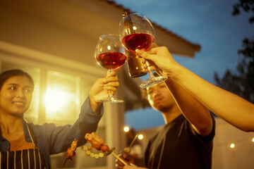 An Asian woman and friends toast with wine during a cozy backyard BBQ. Grilling skewers over charcoal under string lights, they celebrate friendship, joy,  togetherness in a festive outdoor evening