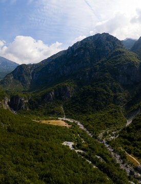 Aerial view of a winding river carves through the verdant valley, embraced by the towering, jagged peaks of the Albanian Alps, Theth, Shkoder County, Albania.