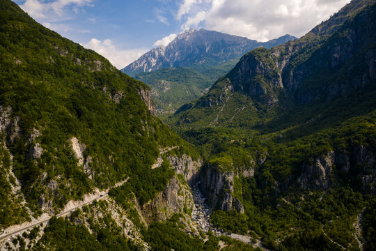 Aerial view of the winding road snaking through the lush, green valley, shadowed by towering, rocky mountains under a bright sky, Theth, Shkoder County, Albania.