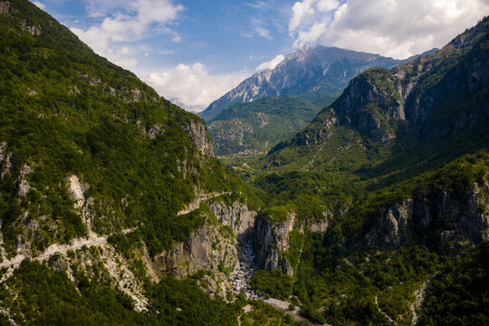 Aerial view of verdant mountains embrace a winding road leading towards a distant, snow-capped peak, nestled in the heart of the valley, Theth, Shkoder County, Albania.