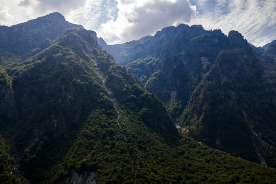 Aerial view of dramatic, rugged peaks ascend into a cloudy sky, their slopes cloaked in deep green forests and exposed rock faces, Theth, Shkoder County, Albania.