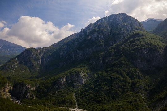 Aerial view of rugged mountains cloaked in verdant forests ascend towards a sky dotted with fluffy white clouds, creating a mesmerizing contrast of textures and tones, Theth, Shkoder County, Albania.