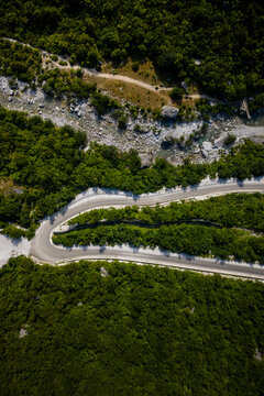 Aerial view of a winding road snaking through the vibrant green forests, contrasted against the stark grey of a rocky riverbed, Theth, Shkoder County, Albania.