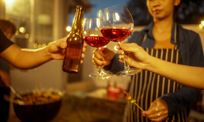 An Asian woman and friends toast with wine during a cozy backyard BBQ. Grilling skewers over charcoal under string lights, they celebrate friendship, joy,  togetherness in a festive outdoor evening