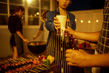 An Asian woman and friends toast with wine during a cozy backyard BBQ. Grilling skewers over charcoal under string lights, they celebrate friendship, joy,  togetherness in a festive outdoor evening