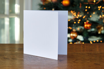 Standing blank empty square greeting card mock up before a blurred christmas tree with copy space....