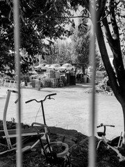 A black and white view through wire fencing, showing a cluttered landfill site with stacked pallets, old equipment, bicycles, and cars mounted on a metal structure for attention