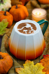 A cup of coffee latte topped with Halloween ghost face draw with cacao powder as latte art with autumn leaves and assorted Jack-Be-Little squash, fall ambiance and candle light.