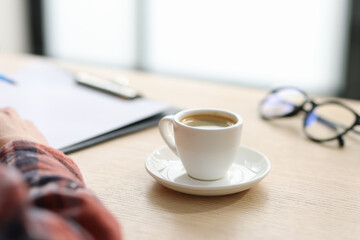 Coffee break. Cup of aromatic drink and woman working at wooden desk indoors, closeup. Space for text