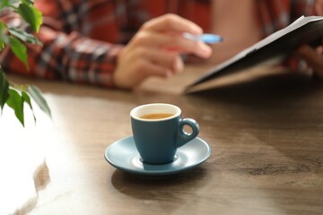 Coffee break. Cup of aromatic drink on wooden table and woman with clipboard indoors, selective focus