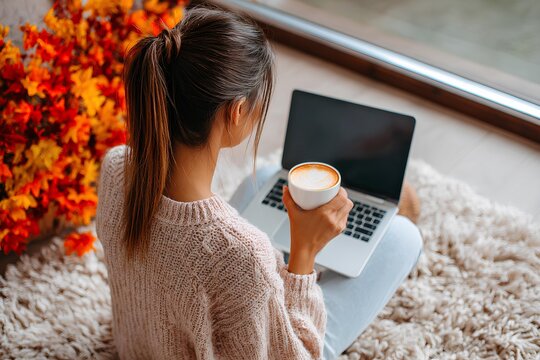 Young woman with long hair, sitting on a cozy rug, working on a laptop while holding a cup of coffee, surrounded by autumn leaves, embodying remote work lifestyle and comfort - Powered by Adobe