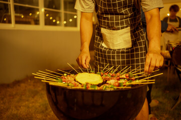 An Asian man wearing a checkered apron carefully arranged skewered meats and vegetables on a round kettle grill. Warm golden lighting highlights the cozy backyard nighttime BBQ atmosphere