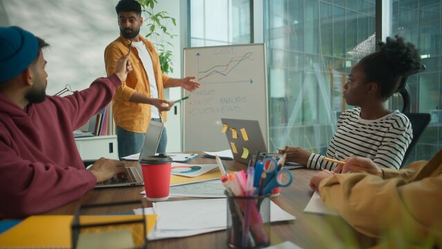 Multicultural informal diversity business team in coworking office listening to male colleague leader presenting financial growth chart on whiteboard discussing startup strategy corporate analytics